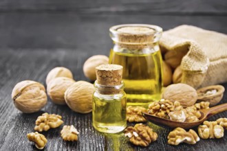 Walnut oil in a glass bottle and a jar, nuts in bag, spoon and on table against dark wooden board
