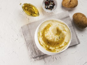 Mashed potatoes in white bowl with cloth napkin, pepper and olive oil on textured concrete white