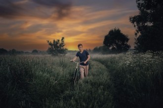 Young man walks with a retro bike along a field road, vintage style in the village at sunset.
