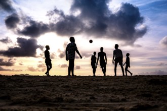 Group of people enjoying a football match on the beach at sunset, silhouettes against a backdrop of
