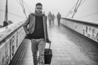 Trendy handsome man walking across a bridge on a misty cold day pulling a suitcase behind him as he