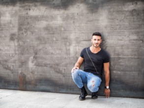 Handsome young man standing against concrete wall, looking at camera, wearing black t-shirt and