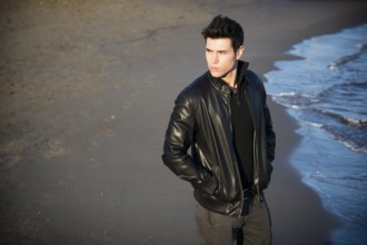 Handsome fashionable young man at the seaside on the beach with his black leather jacket looking