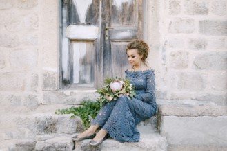 A bride in a stylish blue dress with a bouquet in her hands sits on the steps near an old wooden