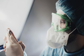 A doctor in green goggles examines a vial while wearing a mask
