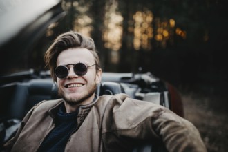 Man smiling broadly in a convertible, wearing sunglasses near a forest at sunset
