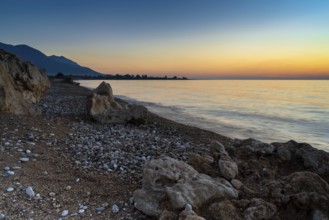 A peaceful sunset over the ocean with silhouette of mountains and pebble beach iwth rock boulders