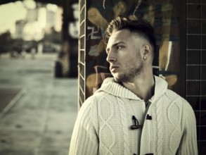Handsome young man in city setting, with industrial site around him, looking away, serious