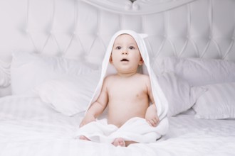 Adorable newborn baby boy with brown eyes sitting wrapped in a white towel with ears in light bed.