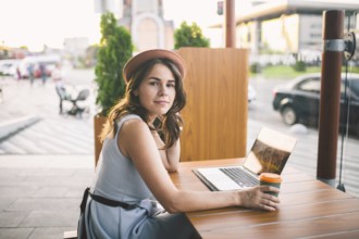 Beautiful young caucasian woman sitting on a terrace in a cafe in summer at a wooden table in a hat