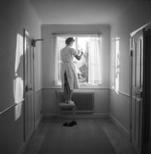 In the 1950s. A woman is seen at a window in a corridor standing barefoot on a chair to hang the