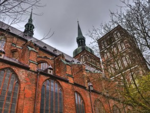 Gothic brick church with towers and large windows under a cloudy sky, surrounded by trees,