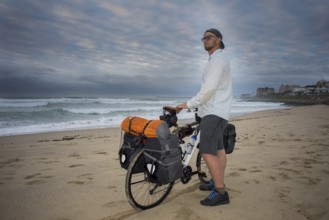 A long distance adventure cyclist stands with his bicycle in hands on the beach while looking at