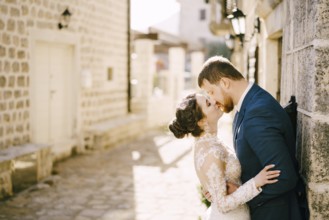 Groom kisses bride near the gray stone wall of the old house.