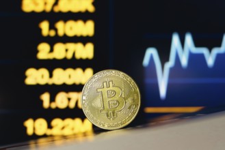 In this photo illustration, a Bitcoin coin is seen on the table and in the background, a stock