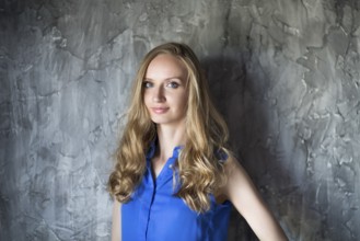 Portrait of the blonde woman in blue blouse staying in front of dark wall and keep smiling