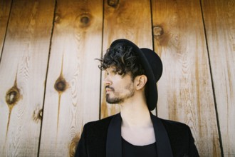 Portrait in profile of a man in a black hat and jacket against a wooden wall.
