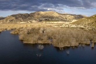 View of a dark blue lake with dry yellow brush and semi-desert mountain and hill landscape behind