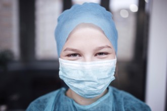 A person in blue scrubs and a medical mask smiles indoors, indicating a healthcare setting