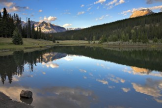 The sun shines tops of distant mountains, clouds above lake and fur-trees on coast