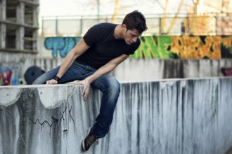 Young man sitting and balancing on dirty wall, looking down, urban environment