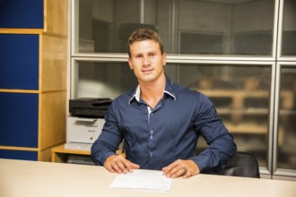 Young man standing, working in an office at the desk with a sheet of paper in front of him, looking