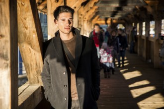 Young dark haired man standing on wooden medieval footbridge in Switzerland while looking away