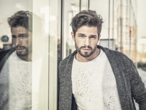 One attractive man in urban environment in city, leaning on glass shop window, wearing cardigan,