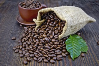 Bag of black coffee beans, brown cup and green leaves on a wooden board background