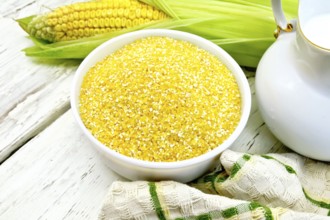 Corn grits in a white bowl, cobs, a jug of milk and a napkin on the background light wooden boards
