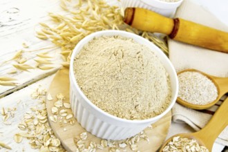 Oat flour in white bowl, oatmeal and bran in wooden spoon, stalks of oats, a rolling pin and a