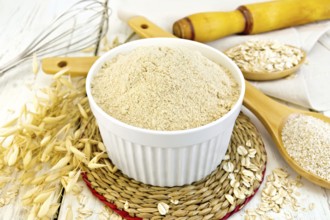 Oat flour in white bowl, oatmeal and bran in wooden spoon, stalks of oats on the background of