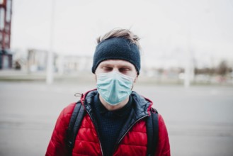 Man in a red jacket and face mask outdoors, with city buildings in the background