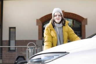 Woman in yellow jacket smiles friendly, at charging station with electric car, environmentally