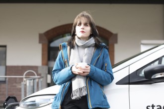 Woman with eyes closed holding a cup and leaning against a charging electric car, environmentally
