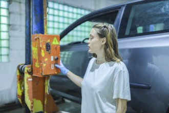 A woman in a workshop thoughtfully examines a switch on a car lift, partially obscured by the car