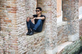 Attractive young man sitting against brick wall, looking in front of himself, wearing sunglasses
