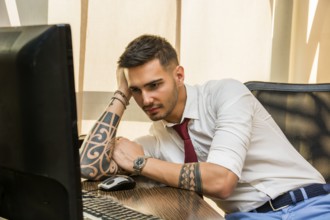 Tired bored young businessman sitting at his desk in front of his computer yawning, holding his
