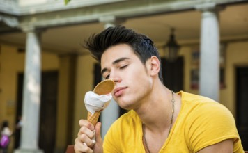 Funny attractive young man sitting, wearing yellow T-shirt while eating a tasty vanilla ice cream