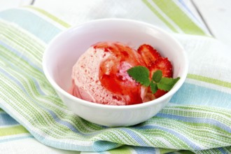 Strawberry ice cream in a white bowl with strawberries and strawberry syrup on a striped linen