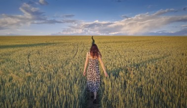 Carefree woman walking a pathway across a green wheat field. Idyllic countryside summer scenery,