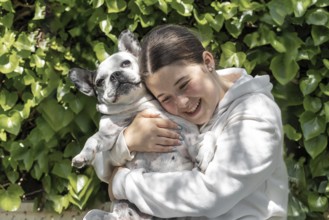 A girl in a white sweatshirt smiling and hugging a French Bulldog in a garden with lush green