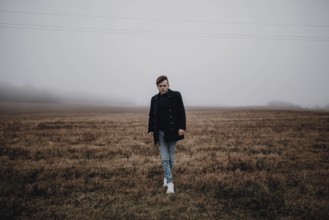 A man in a coat stands in a foggy field, evoking a moody, autumnal atmosphere