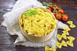 Farfalle pasta with basil in a bark box on a burlap napkin, tomatoes against dark wooden board