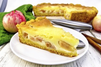 Piece of apple pie with sour cream sauce in a white plate, cinnamon, fruit and napkin on a wooden