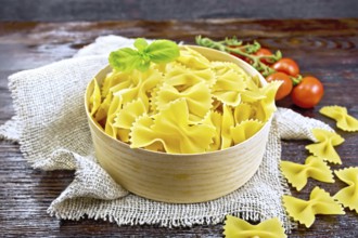 Farfalle pasta with basil in bark box on a burlap napkin, tomatoes on wooden board background