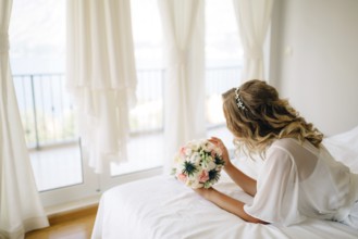 A bride in a gentle peignoir with a bouquet in her hands laying on the bed by the window with white