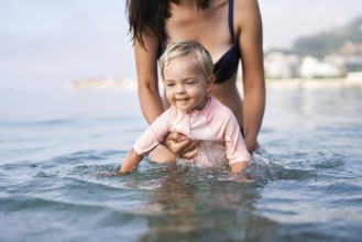 Little girl bathes in shallow water supported by her mom.