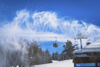 Snow cannon spreading fresh snow powder on a ski slope. Skiers riding up on chair lift. Winter ski