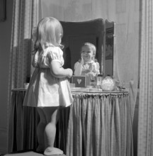 In the 1950s. A little girl is standing in front of her mothers makeup table and mirror and puts on